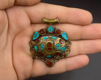 May include: An ornate, antique pendant with a brass-colored metal base. The pendant is adorned with turquoise and red-colored stones, arranged in a symmetrical pattern. The design features intricate swirls and a loop at the top for a chain.