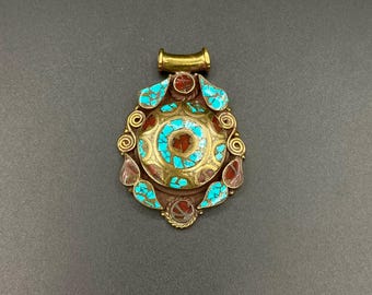 May include: Ornate pendant with a gold-colored bail and a central design featuring a red and turquoise circular pattern. The pendant is adorned with turquoise and red-toned stones, and gold-colored filigree. The pendant is set against a dark gray background.