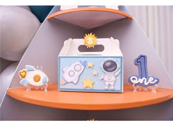May include: A blue and white cardboard box with a handle, decorated with a rocket, astronaut, stars, and a sun. The box is on a shelf with a blue and white cookie that says "one" and a rocket cookie.