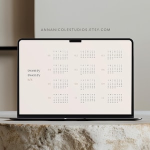 Puede incluir: Un portátil que muestra un calendario, colocado sobre una superficie de piedra clara y texturizada. La pantalla muestra un diseño de calendario anual con una combinación de colores neutros. El fondo presenta estantes con cerámica y una pared neutra.