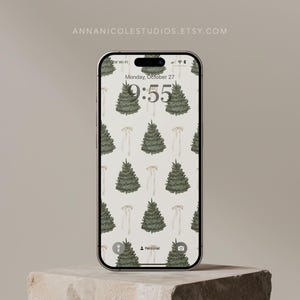 May include: A smartphone displays a patterned wallpaper featuring green Christmas trees and white floral elements on a cream background. The time is 9:55 on Monday, October 27. The phone is on a stone block. The website annnicolestudios.etsy.com is visible.