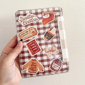 May include: A clear phone case with a red and white checkered pattern, decorated with fall-themed stickers. Stickers include text that says "Sweater Weather", "Cozy Girl Bookclub", "Cozy Vibes Only", "Team Stay Inside & Read", and "Pumpkin Spice".