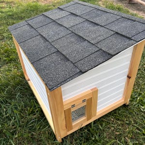 Pode incluir: Uma casinha de madeira para animais de estima&ccedil;&atilde;o com telhado de telhas cinzentas. A casa tem revestimento branco com ranhuras verticais e uma pequena porta com uma aba transparente. A estrutura &eacute; de madeira natural.