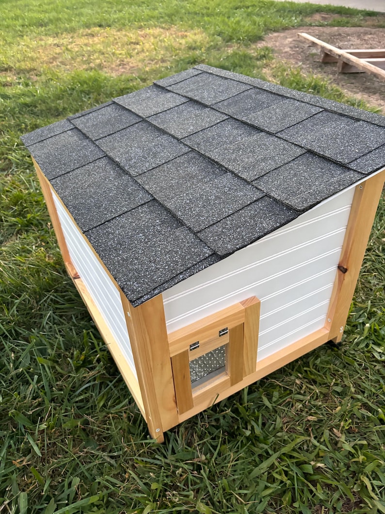 Pode incluir: Uma casinha de madeira para animais de estima&ccedil;&atilde;o com telhado de telhas cinzentas. A casa tem revestimento branco com ranhuras verticais e uma pequena porta com uma aba transparente. A estrutura &eacute; de madeira natural.