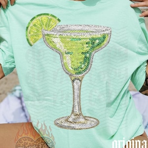 May include: A light green t-shirt featuring a sequined margarita glass design with a lime wedge. The shirt has short sleeves and a relaxed fit. The words "original designer" are printed on the shirt.