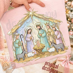 May include: A light pink sweatshirt featuring a printed nativity scene illustration with gold glitter accents. The design depicts figures in pastel robes, a manger with a baby, two donkeys, and two sheep under a golden star-topped stable roof.