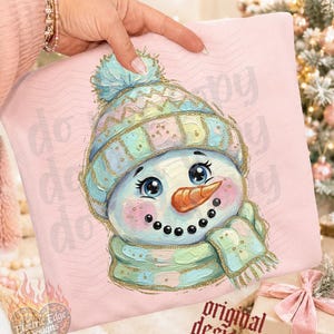 May include: A light pink sweatshirt featuring a central illustration of a snowman wearing a pastel-colored knit hat with a pom-pom and a matching scarf. The snowman has a bright orange carrot nose, rosy cheeks, and large blue eyes.