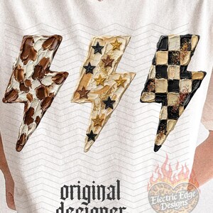 May include: Cream-colored t-shirt featuring three lightning bolt designs with unique patterns: cow print, stars, and a black and tan checkerboard. The shirt has the text "original designer" in a gothic font.