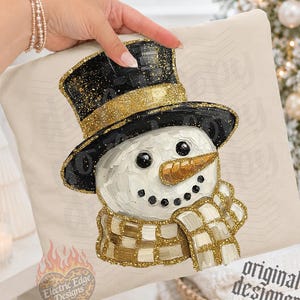May include: A cream-colored fabric item featuring a textured, painted-style illustration of a snowman wearing a black top hat with a gold glitter band and a matching gold and white checkered scarf. The snowman has a gold carrot nose and black eyes.