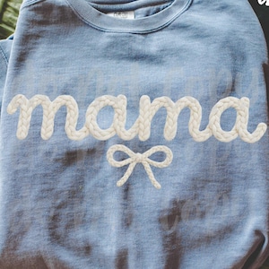 Puede incluir: Sudadera azul claro con la palabra "mama" en blanco, con letras tipo cuerda y un lazo blanco. Las palabras "original designer" están escritas en blanco en la esquina superior derecha. Una bufanda verde está en el fondo.