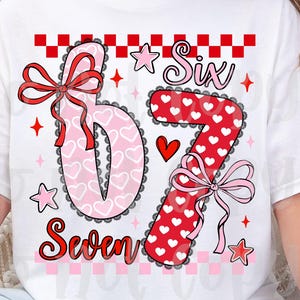 May include: White t-shirt with a colorful graphic design featuring the numbers "6" and "7" in a heart and polka dot pattern, with the words "Six" and "Seven". The design includes bows, stars, and a heart.