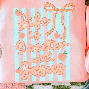 May include: A coral-colored sweatshirt with the text "Life is Sweeter with Jesus" in a playful font, accented with peaches and a bow. The design is set against light blue stripes. The sweatshirt is displayed on a checkered green and white surface.
