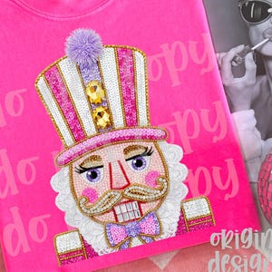 May include: A vibrant pink sweatshirt with a detailed nutcracker design. The nutcracker has a sequined hat, a purple pom-pom, and a bow tie. The words "original designer" are at the bottom right.