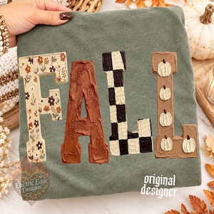 May include: An olive green sweatshirt featuring the word FALL in large block letters. The F has a floral pattern, the A is textured brown, the L is a black and white checkerboard, and the L features small white pumpkins on a brown background.