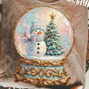 May include: A taupe t-shirt featuring a graphic of a snow globe containing a snowman wearing a blue hat and scarf next to a Christmas tree. The snow globe has an ornate gold base and the text original designer is printed in white script.