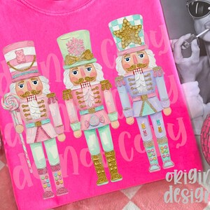 May include: A bright pink t-shirt featuring three colorful nutcracker illustrations. Each nutcracker has a unique design, with gold accents and pastel colors. The text "Candy Candy" is printed on the shirt. The brand label "Comfort Colors" is visible.