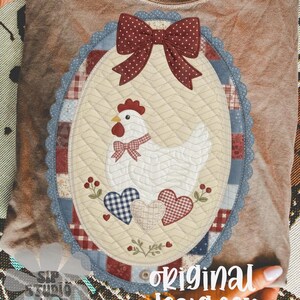 May include: A beige t-shirt with a quilted oval design featuring a white chicken, red bow, and hearts. The design is framed by a blue and red border. The text "original designer" is visible.