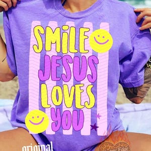May include: A purple t-shirt with the text "SMILE JESUS LOVES YOU" in yellow and pink. The design includes smiley faces and stars. The shirt is a short-sleeved, casual style.