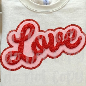 May include: Cream-colored sweatshirt with the word "Love" in a red, glittery, bubble letter design. The word is outlined in a lighter pink and red. The sleeves are rolled up.