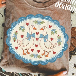 May include: A brown t-shirt featuring a whimsical design of two geese with blue bow ties, surrounded by hearts and floral accents within a blue scalloped oval. The words "original designer" are printed on the shirt.