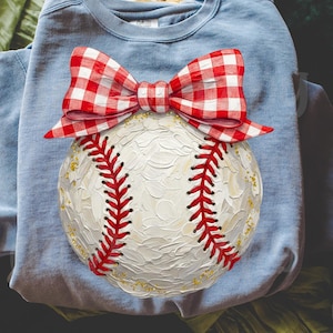 Peut inclure: Un sweat-shirt bleu clair avec un motif de baseball. La balle est blanche avec des coutures rouges et un n&oelig;ud vichy rouge et blanc. Le baseball a des d&eacute;tails dor&eacute;s.
