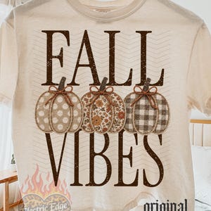 Könnte beinhalten: Ein cremefarbenes Kurzarm-T-Shirt mit dem Schriftzug FALL VIBES in dunkelbraunen Buchstaben. Unter dem Text befinden sich drei Kürbisillustrationen mit verschiedenen Mustern: Punkte, Blumen und Karomuster, jeweils mit einer braunen Bindfaden-Schleife.