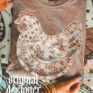 Könnte beinhalten: Ein braunes T-Shirt mit einem floralen Hühner-Applikation. Das Hühnerdesign zeigt eine Mischung aus floralen Mustern in Rosa, Creme und Braun. Die Worte "original designer" sind in einer gotischen Schriftart gedruckt. Ein herzförmiger Aufkleber mit dem Text "Electric Edge Designs" ist ebenfalls sichtbar.