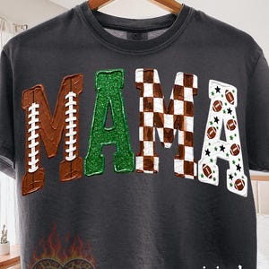 May include: A charcoal gray t-shirt featuring the word MAMA in large letters. The M is brown with football laces, the A is green glitter, the M is a brown and white checkerboard pattern, and the A is white with footballs and stars.