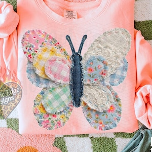 May include: A coral-colored sweatshirt features a patchwork butterfly design with floral and patterned wings. The butterfly is centered on the chest. The words "original designer" are printed on the bottom right of the image.