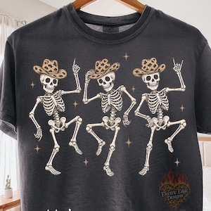 May include: A charcoal gray short-sleeve t-shirt featuring a graphic of three dancing skeletons wearing leopard print cowboy hats. The design includes small star accents and the text "original designer" in white, along with a heart-shaped logo.