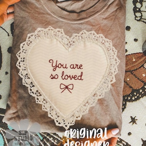 May include: A taupe t-shirt with a heart-shaped appliqu&eacute; embroidered with the words "You are so loved" and a bow. The heart is trimmed with lace. The shirt is folded and displayed on a patterned surface, with the words "original designer" visible.