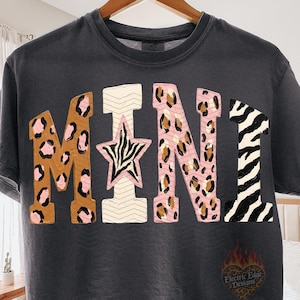 May include: A charcoal gray t-shirt featuring the word MINI in large block letters. The letters have various patterns including brown and pink leopard print, a pink star with zebra stripes, cream chevron lines, and black and white zebra stripes.