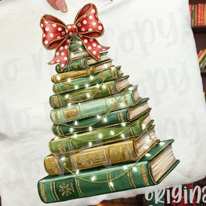 May include: White pillow with a Christmas tree design made of stacked books. The books are green and gold, adorned with string lights and topped with a red bow with white polka dots. The pillow is held by a hand.