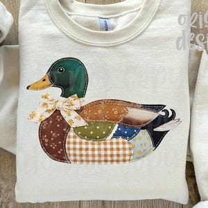 May include: Cream-colored sweatshirt featuring a patchwork duck design. The duck has a green head, yellow beak, and a bow. The body is made of various fabric patterns, including brown, blue, and gingham. The sleeves are rolled up.