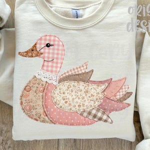 May include: Cream-colored sweatshirt featuring a patchwork duck design. The duck has a pink gingham head, a brown beak, and a floral patterned body. The wings are made of various pink and brown fabrics. The sleeves are a solid cream color.