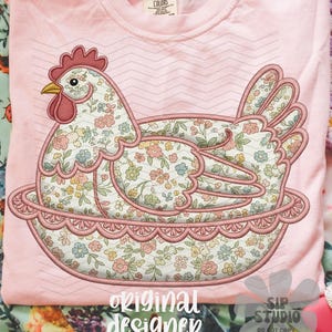 May include: A light pink t-shirt featuring a floral hen design. The hen is embroidered with a colorful floral pattern and sits in a decorative nest. The text "original designer" is printed at the bottom.