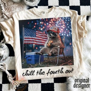 K&ouml;nnte beinhalten: Cremefarbenes T-Shirt mit einem Waschb&auml;ren, der eine Sonnenbrille und einen roten Hut mit der Aufschrift "America" tr&auml;gt und einen Hotdog h&auml;lt. Der Waschb&auml;r sitzt in einem Stuhl vor einer amerikanischen Flagge und Feuerwerk. Der Text "chill the fourth out" steht darunter.