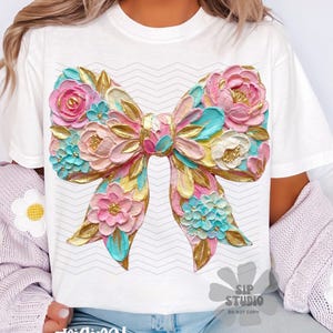 May include: White t-shirt featuring a large, colorful bow design with floral accents. The bow is rendered in shades of pink, blue, gold, and yellow, with a textured appearance. The shirt also has the text "original designer" at the bottom.