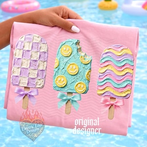 May include: A light pink t-shirt featuring three popsicle graphics with textured frosting. The left popsicle has a purple and white checkerboard pattern, the middle is light blue with yellow smiley faces, and the right has wavy stripes.