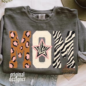 Puede incluir: Una sudadera gris carbón de cuello redondo con la palabra MOM en letras grandes. La M es marrón con manchas de leopardo rosas, la O es crema con una estrella de estampado de cebra con borde rosa en el centro, y la última M tiene rayas de cebra negras.