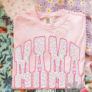 Op de afbeelding: Een lichtroze Comfort Colors sweatshirt met het woord "MAMA" in grote, bloemrijke letters met roze omtrek. De woorden "original designer" staan onderaan. Een gehaakte madeliefjesdeken is op de achtergrond.