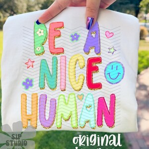 May include: White t-shirt with the phrase "BE A NICE HUMAN" in colorful, glittery letters. The design includes flowers, stars, a heart, and a smiling face. The words "SIP STUDIO" and "original designer" are also printed on the shirt.