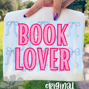 May include: White t-shirt with the words "BOOK LOVER" in pink, set against a light blue striped background with bow accents. The shirt also features the text "original designer".