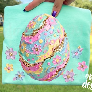 May include: A mint green sweatshirt featuring a large, textured Easter egg design in pastel colors with gold glitter accents and floral embellishments. The words "original designer" are printed in white.