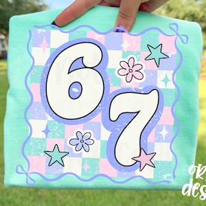 May include: A light green t-shirt with a colorful graphic featuring the numbers "6" and "7" in a retro font. The graphic includes stars, flowers, and a checkered pattern in pastel colors. The text "original designer" is in the bottom right corner.