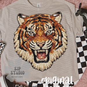 May include: Beige t-shirt featuring a detailed tiger face design. The tiger's face is embellished with sequins in shades of gold, orange, and black. The shirt is paired with black and white checkered accessories and a black sneaker.