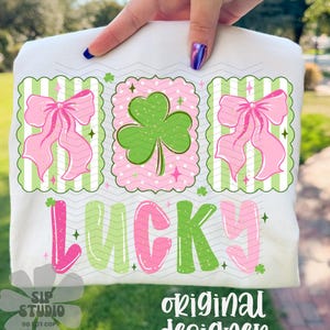 May include: White t-shirt featuring a St. Patrick's Day design. The design includes pink bows, a green shamrock, and the word "LUCKY" in pink and green. The shirt is held up by a person.
