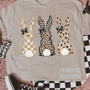 May include: A beige t-shirt featuring three bunny designs with checkered, leopard, and spotted patterns. Each bunny has a black bow and a white floral accent. The text "original designer" is printed on the shirt.