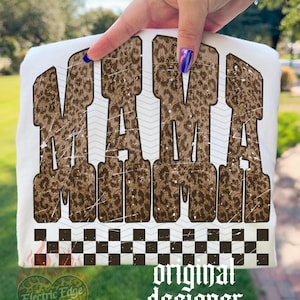 May include: White t-shirt with the word "MAMA" in brown leopard print lettering. The shirt has a black and white checkered pattern at the bottom and the words "original designer" in a gothic font.