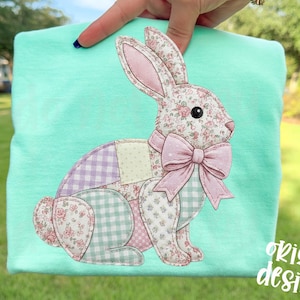 Puede incluir: Una camiseta verde azulado claro con un diseño de conejo de patchwork. El conejo está hecho de patrones florales y de cuadros en rosa, morado y verde. Tiene un lazo y orejas rosas. El texto "original designer" está escrito en blanco.